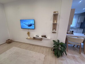 A modern living room with a flat-screen TV mounted on the wall, a small white shelf with decorative items, a potted plant, and a dining area with a table and chairs. - New Listing (Chéraga)
