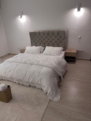 A bedroom with a tufted headboard, white bedding, a wooden nightstand, and two wall-mounted lights. - New Listing (Chéraga)