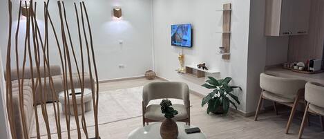 A modern living room with a sofa, a glass coffee table, a TV mounted on the wall, and a kitchen area with wooden cabinets.
