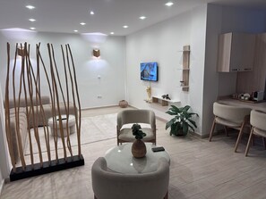 A modern living room with a sofa, a glass coffee table, a TV mounted on the wall, and a kitchen area with wooden cabinets. - New Listing (Chéraga)