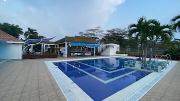 Una piscina con borde de baldosas, un área techada con asientos y palmeras al fondo.