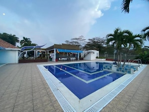 A swimming pool with a tiled deck, a covered area with seating, and palm trees in the background. - Hotel Campestre Serrania de Manacacias (SAN MARTÍN)