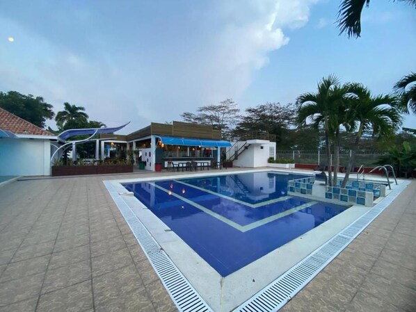 A swimming pool with a tiled deck, a covered area with seating, and palm trees in the background.