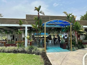 An outdoor seating area with tables and chairs, surrounded by greenery and a covered structure.