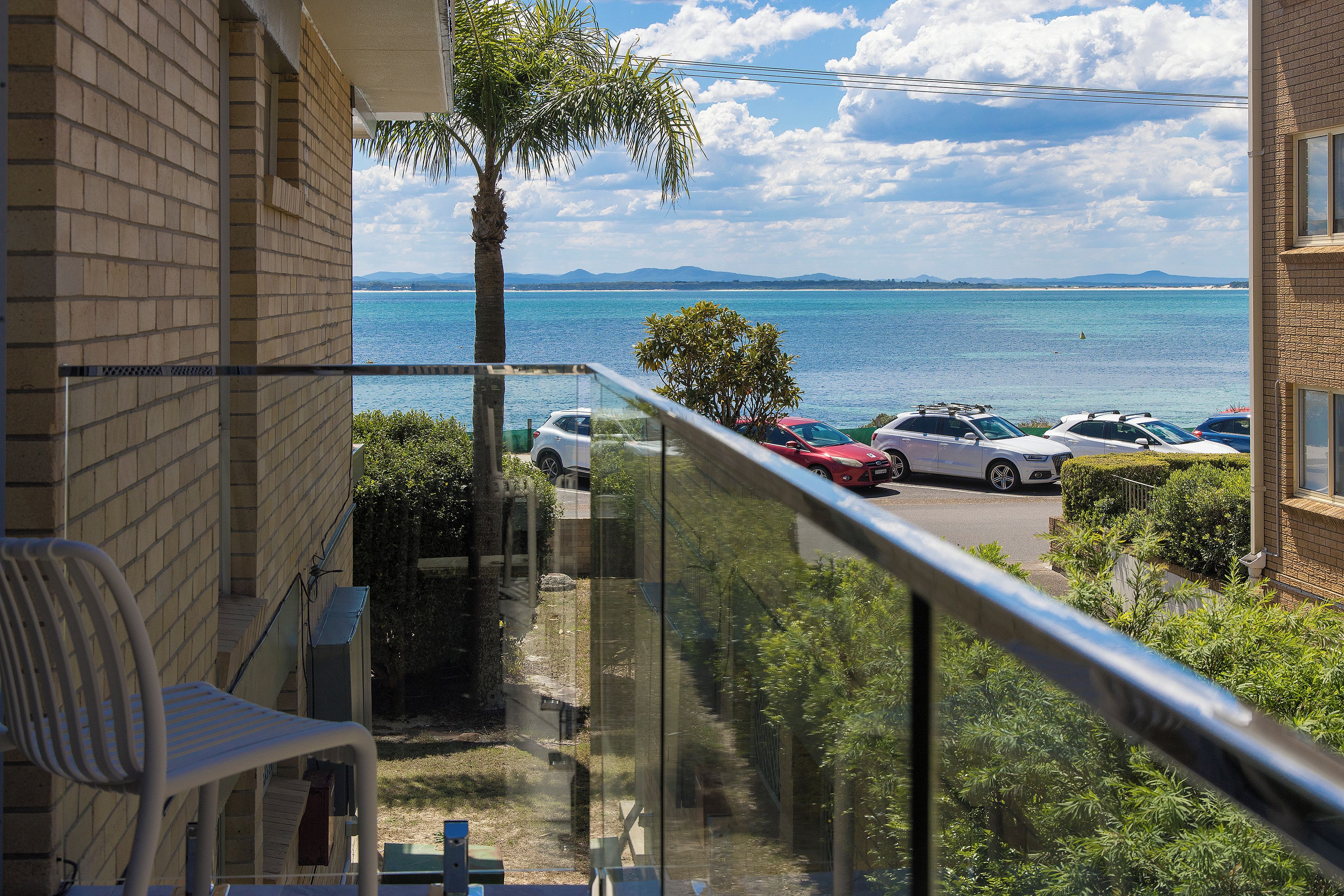 A balcony with a view of the sea, parked cars, and a clear sky.