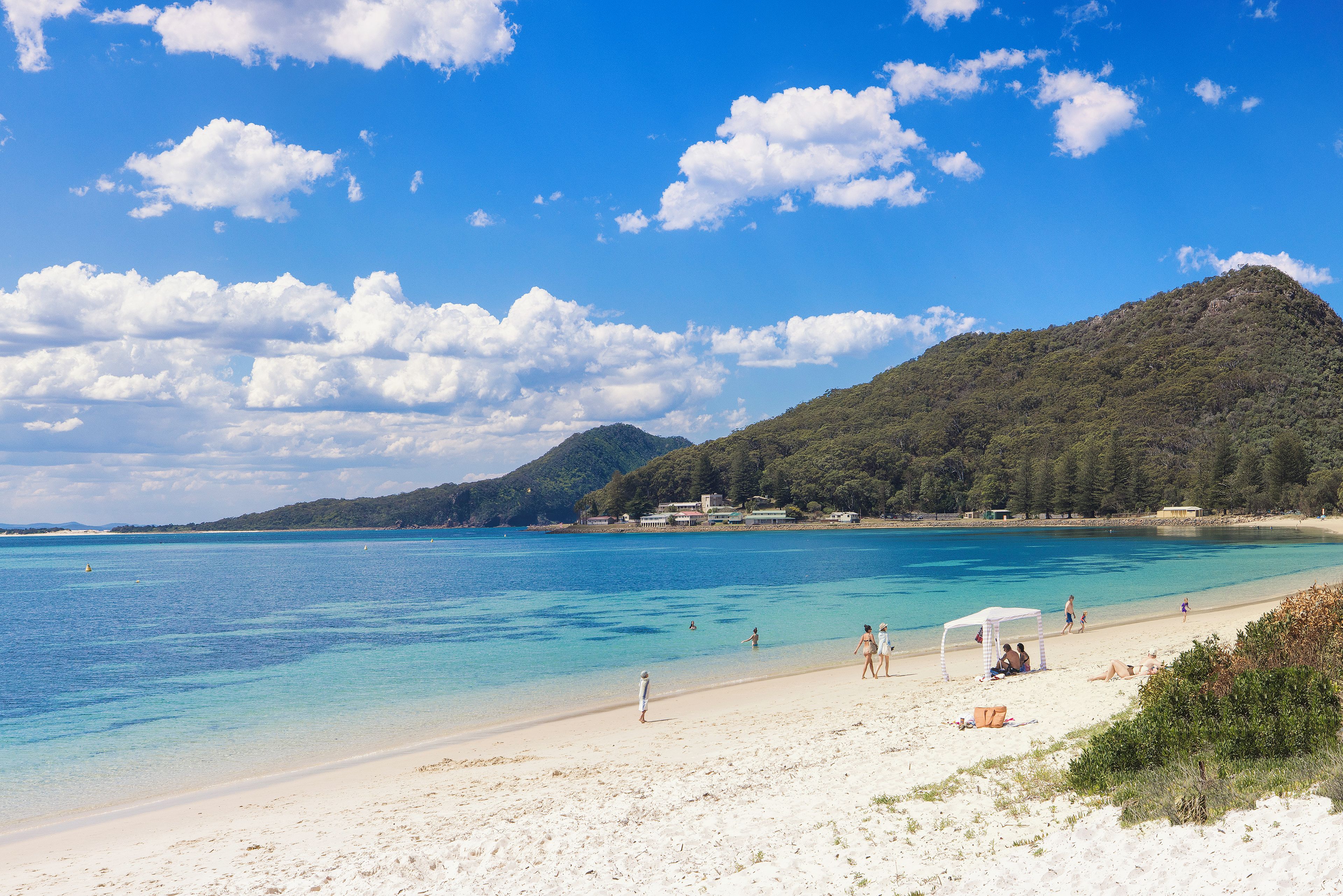 Ein Strand mit klarem, blauem Wasser, sandigem Ufer und einem Berg im Hintergrund.