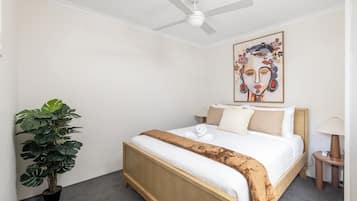 A hotel room with a bed, a ceiling fan, a large painting, a potted plant, and a bedside table with a lamp.