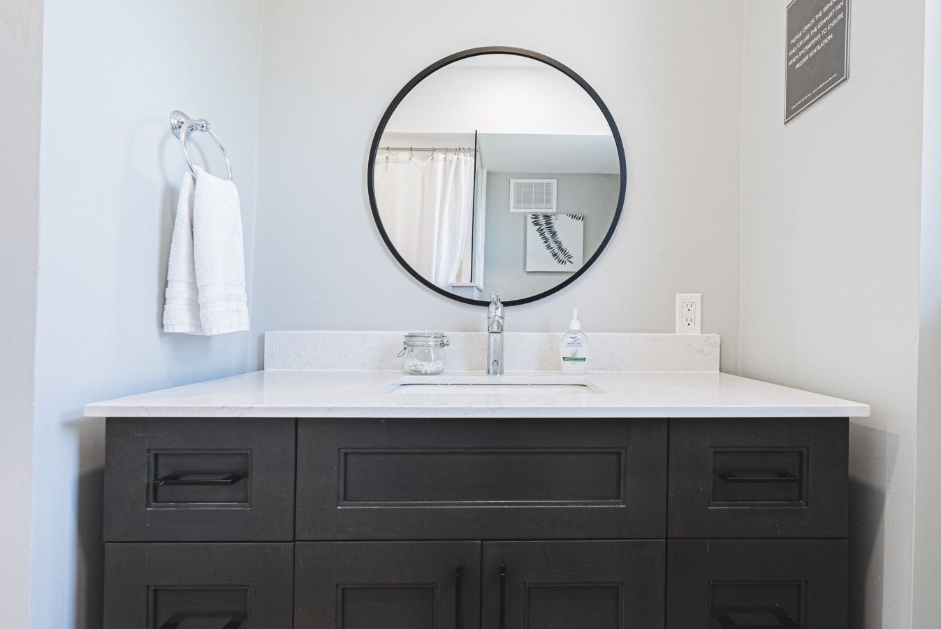 Une salle de bain avec un lavabo blanc, des meubles sombres, un miroir rond et une serviette accrochée au mur.