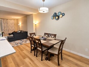 A dining area with a wooden table and chairs, a sofa in the background, and a wall-mounted light fixture. - Modern 3BR/2.5BA Home | Free Parking, WiFi, Full Kitchen, Laundry & Deck (Regina)
