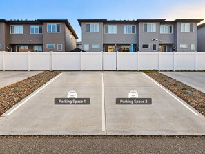 Un espace de stationnement avec deux places délimitées, une rangée d’immeubles d’appartements modernes et une clôture blanche.