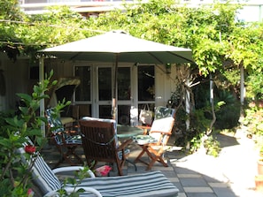 Een terras buiten met een overdekte zithoek, inclusief een tafel en stoelen, omringd door groen.