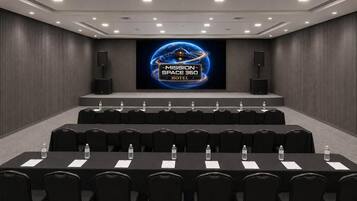 A conference room with a large screen displaying 'MISSION SPACE 360 HOTEL' logo, multiple rows of black chairs, and water bottles on the tables.