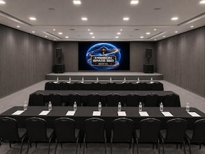 Un salón de conferencias con una gran pantalla mostrando el logotipo 'MISSION SPACE 360 HOTEL', varias filas de sillas negras y botellas de agua sobre las mesas.