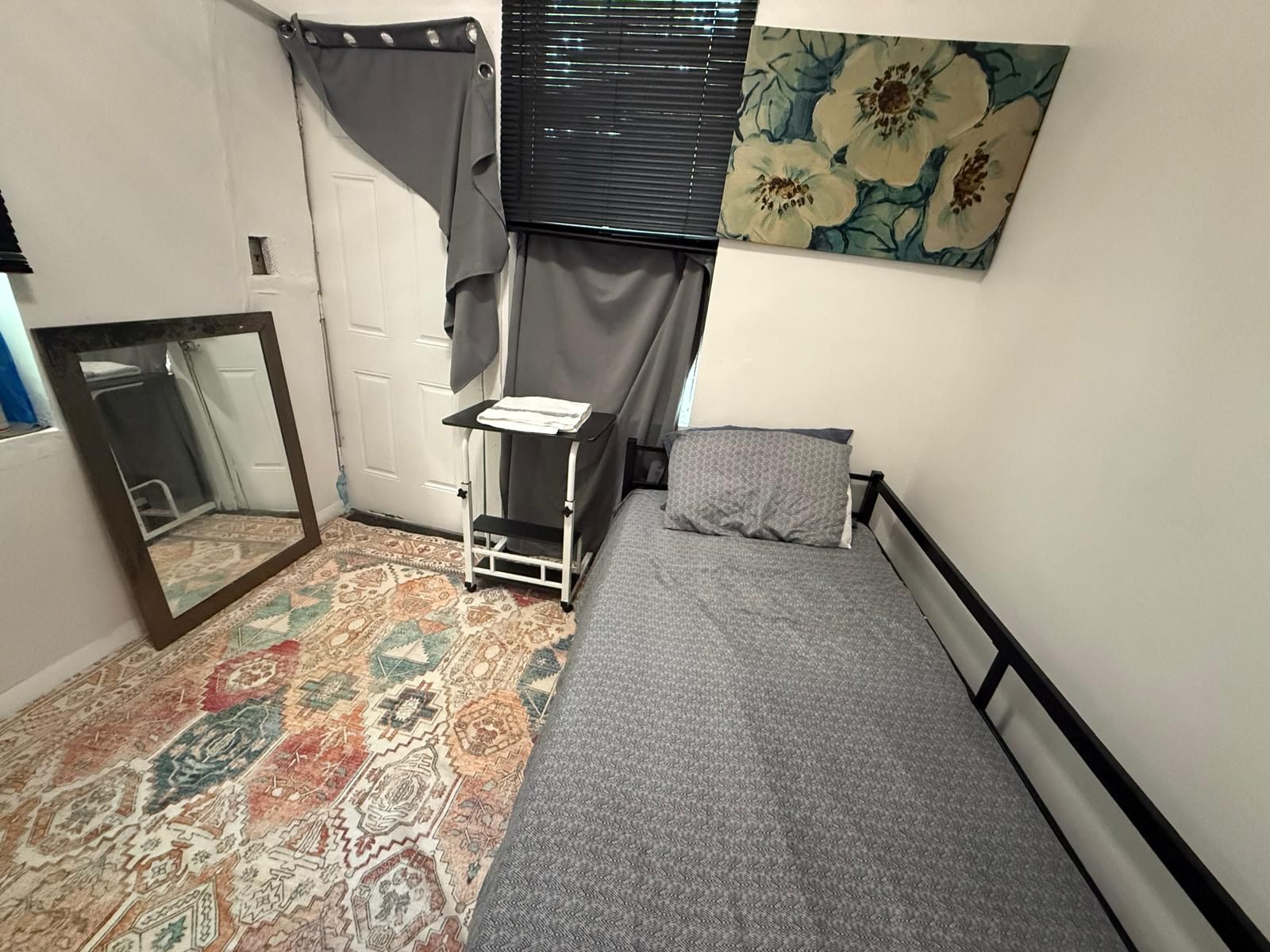 A small bedroom with a bed, a nightstand, a mirror, a window with curtains, and a floral painting on the wall.