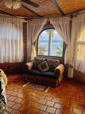 A cozy room with a brick wall, a cushioned chair with decorative pillows, a view of the sea, and a tiled floor. - New Listing (San Juan Tecomatlan)