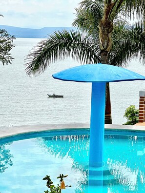 Een zwembad met een blauwe parasol, palmbomen en uitzicht op de zee.