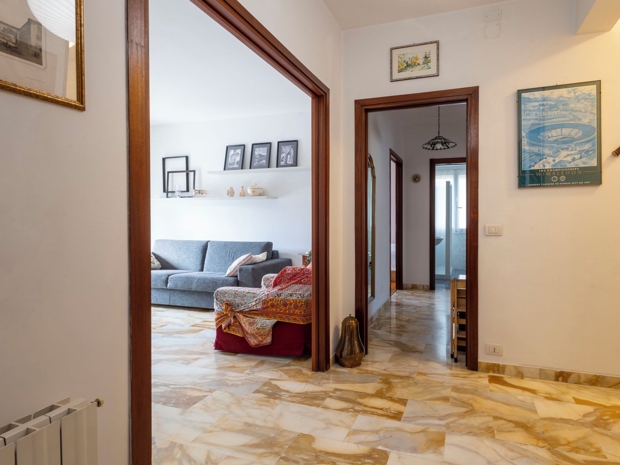 A hallway with a sofa, a patterned ottoman, and a mirror reflecting a room with a window.