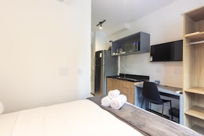 A compact hotel room with a bed, a desk with a chair, a kitchenette, and a TV mounted on the wall. - Studio in Condo. w/ Pool and Terrace Oju0208 (São Paulo)