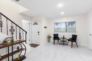 Ein helles, modernes Interieur mit Treppe, einer Tür, zwei Stühlen, einem kleinen Tisch und einem Fenster mit Jalousien.