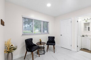 A room with two black upholstered chairs, a small round table, a vase with flowers, and a window with blinds. - Charming 3-bedroom house in tranquil Dade City with AC (Dade City)