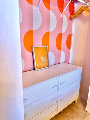 Ein Zimmer mit einer weißen Sitzbank an einer Wand mit geometrischem Muster, einem gerahmten Bild auf der Sitzbank und einer weißen Kommode mit Schubladen.