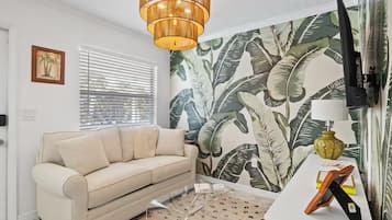 Un salon avec un canapé beige, une table basse en verre transparent, un téléviseur fixé au mur et un papier peint à motifs.