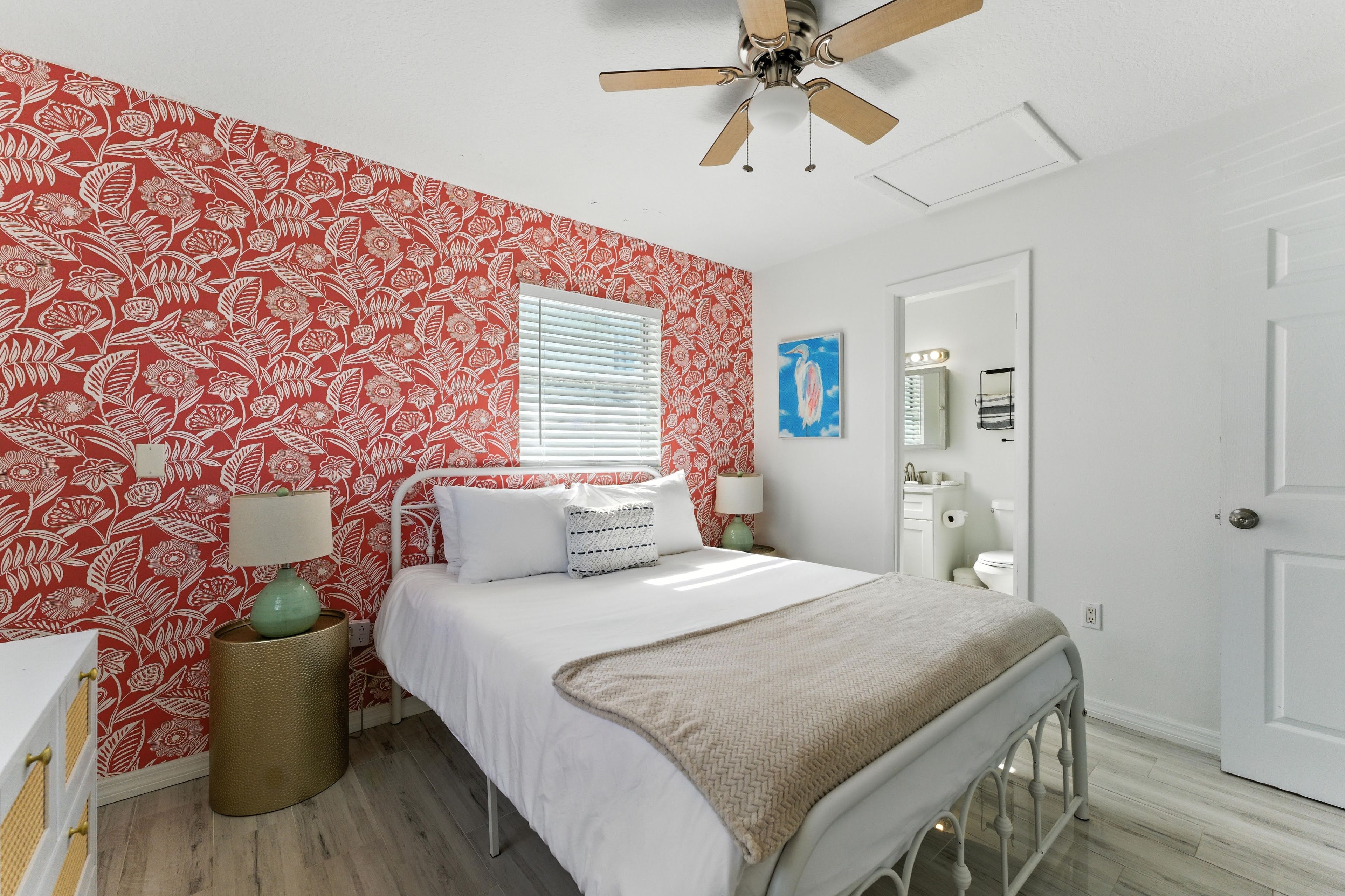 A bedroom with a bed, a bedside table, a lamp, a window with blinds, a ceiling fan, and a door.