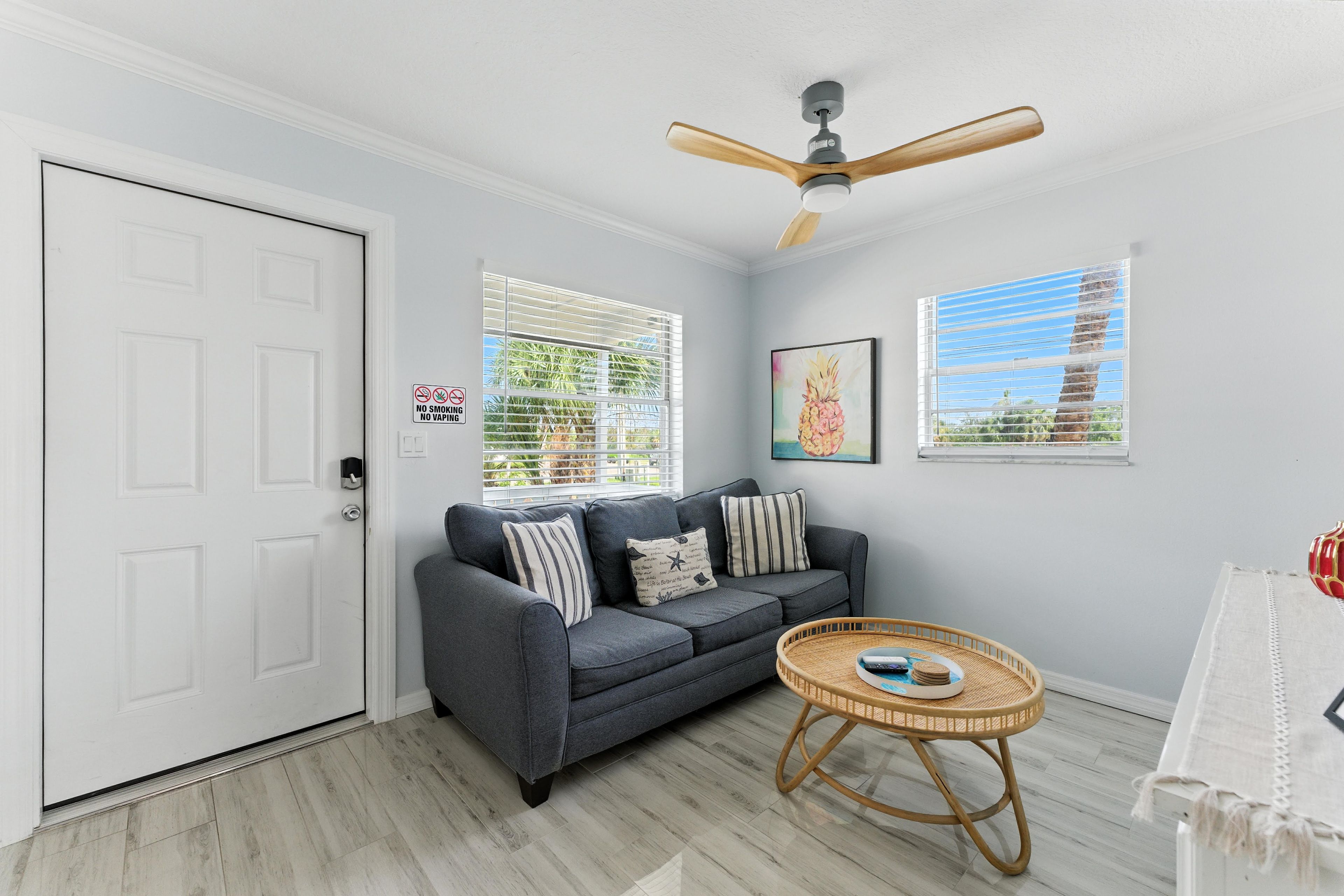 A living room with a grey sofa, a round wicker table, a ceiling fan, and two windows with blinds.