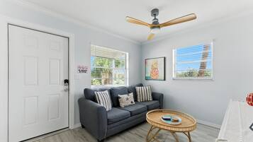 A living room with a grey sofa, a round wicker table, a ceiling fan, and two windows with blinds.