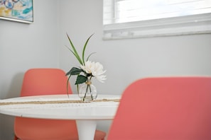 Une table ronde recouverte d’une nappe blanche, un vase avec des fleurs blanches et deux chaises d’une teinte rosée.
