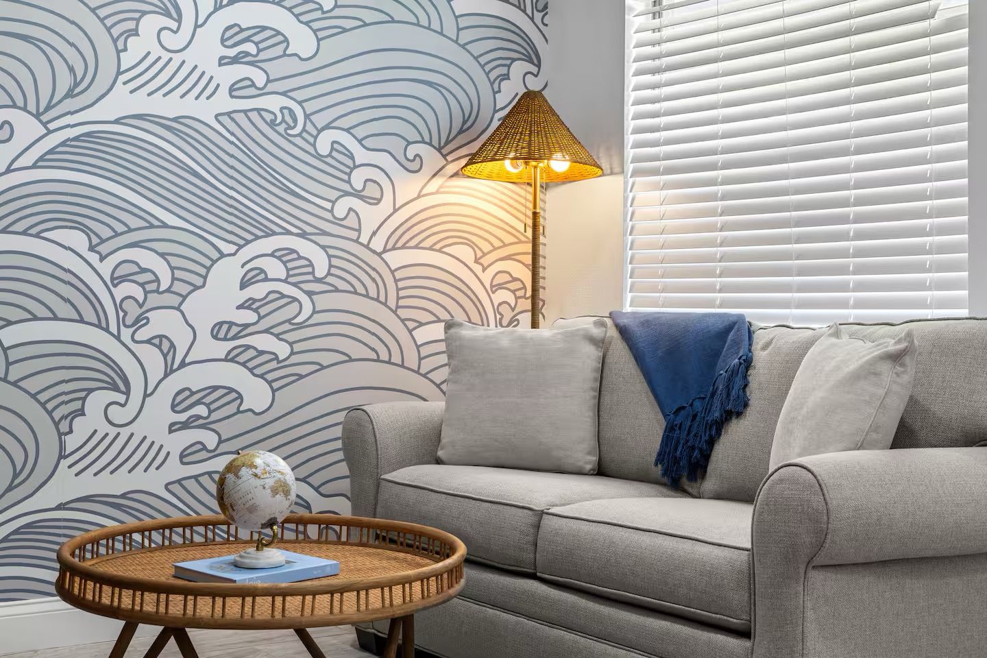 Un salon avec un canapé gris, une table basse ronde en osier sur laquelle reposent un globe terrestre et un livre, un lampadaire et un mur orné d’un motif ondulé.