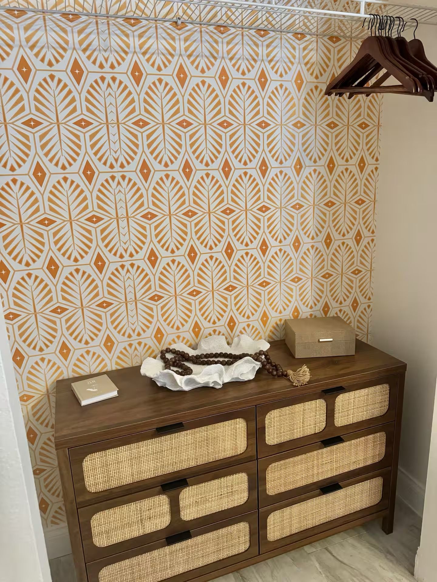A room with a patterned wallpaper, a wooden dresser with woven drawers, and a hanging clothes rack.