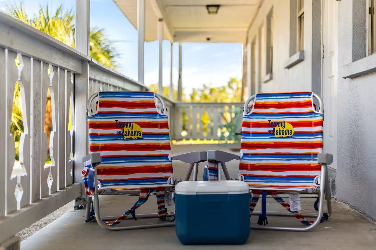 Deux chaises de plage au motif tropical, une glacière bleue et une véranda avec une balustrade.