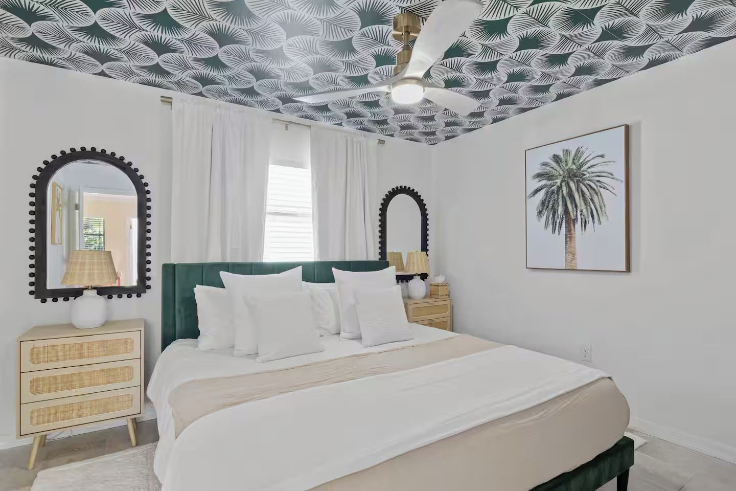 Une chambre à coucher comprenant un lit, deux tables de chevet, un miroir, un ventilateur de plafond et un tableau encadré accroché au mur.