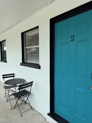 Une porte de couleur turquoise portant le numéro 2, une fenêtre avec des stores et une petite table d’extérieur avec des chaises.