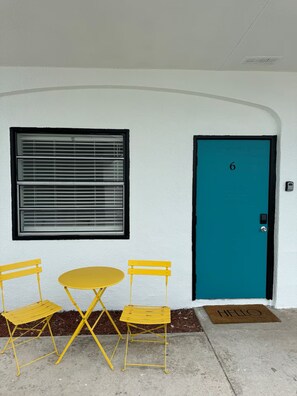 Un bâtiment avec une porte de couleur turquoise, une fenêtre avec des stores et une table et des chaises jaunes disposées à l’extérieur.