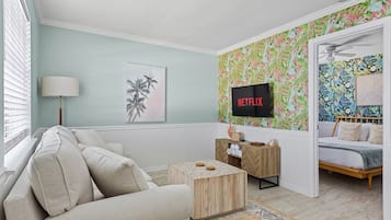Una sala de estar con sofá, mesa de centro, televisor montado en la pared y cama con cabecera.