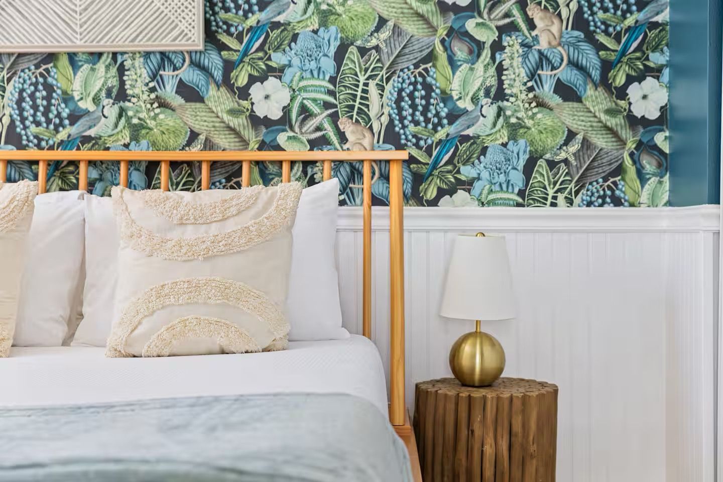 A bed with white bedding and decorative pillows, a wooden headboard, a wall with a floral mural, and a small round wooden nightstand with a gold lamp.