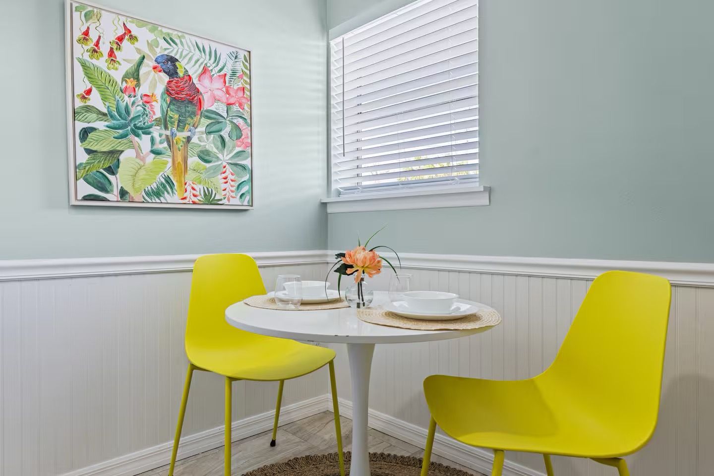 Un comedor con una mesa redonda, dos sillas amarillas, un mantel blanco y un centro de mesa floral.