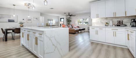 A spacious kitchen with white cabinetry and marble countertops, a dining area with a wooden table, and a living room with a sofa and coffee table.