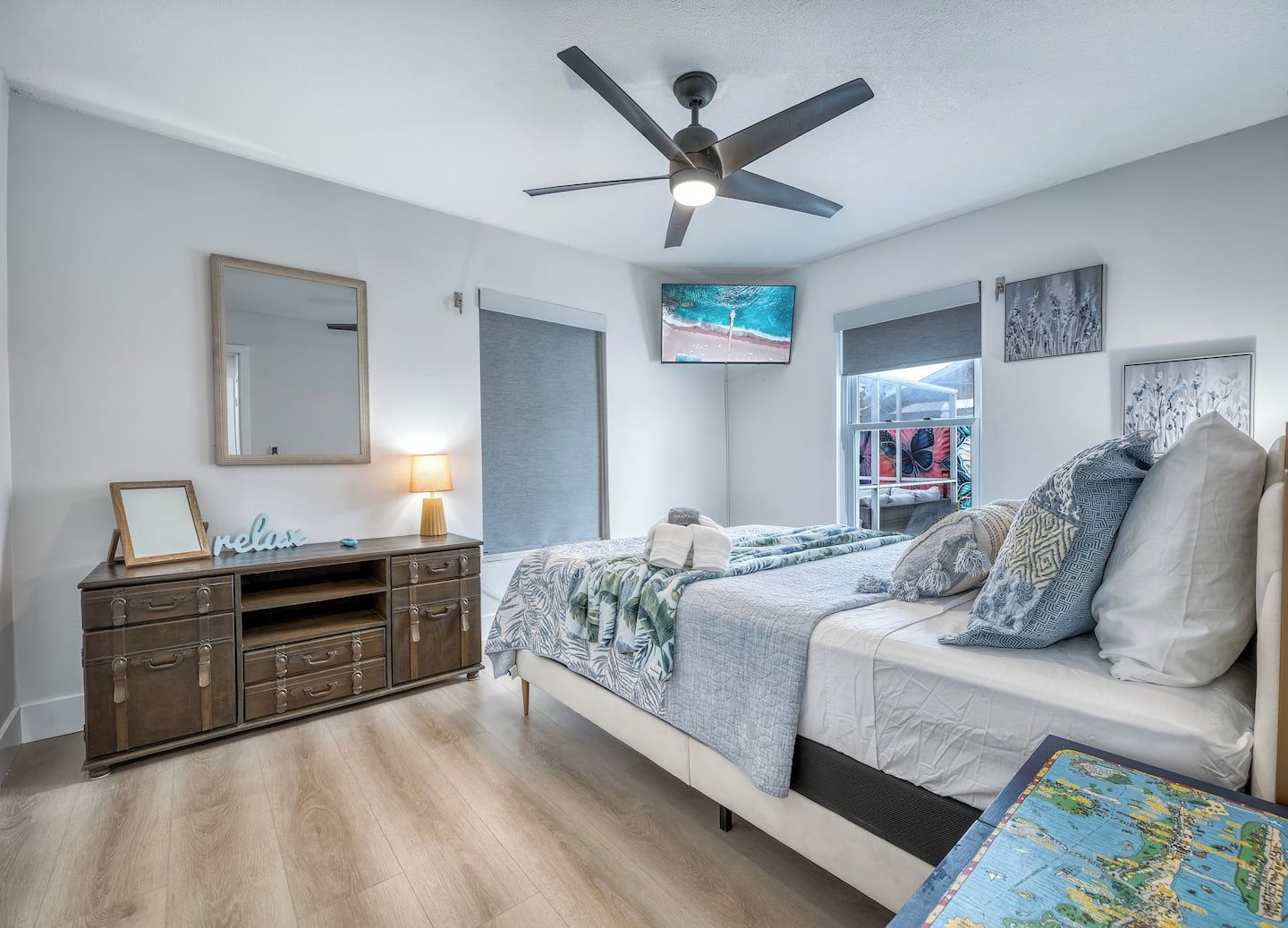 Un dormitorio con cama, cómoda, ventilador de techo, ventana y obras de arte en las paredes.