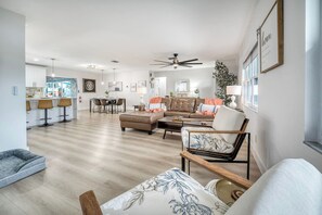 Ein großzügiges Wohnzimmer mit Sofa, Sessel und Hocker, ein Essbereich mit Stühlen, eine Küche im Hintergrund und ein Deckenventilator.