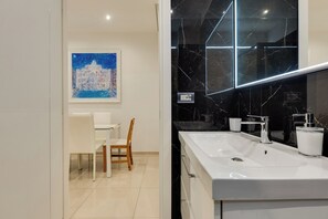 A modern bathroom with a white sink, a mirror with a black marble backsplash, and a wooden chair. - New Listing (Roma)