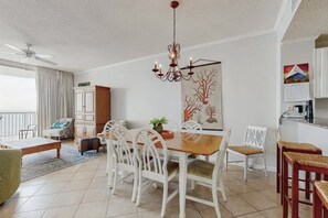 A dining area with a wooden table and white chairs, a chandelier, and a painting of trees on the wall. - Beachfront Resort Condo With Pools, Spa, Club House and Pickleball Court (Gulf Shores)