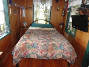 A bedroom with a wooden floor, a bed with a colorful quilt, a window with lace curtains, and a television on the wall. - Lovers Lane Getaway (Wellington North)