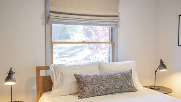 A neatly made bed with white bedding and a patterned pillow, flanked by two bedside lamps, in a room with a window and a plain wall.