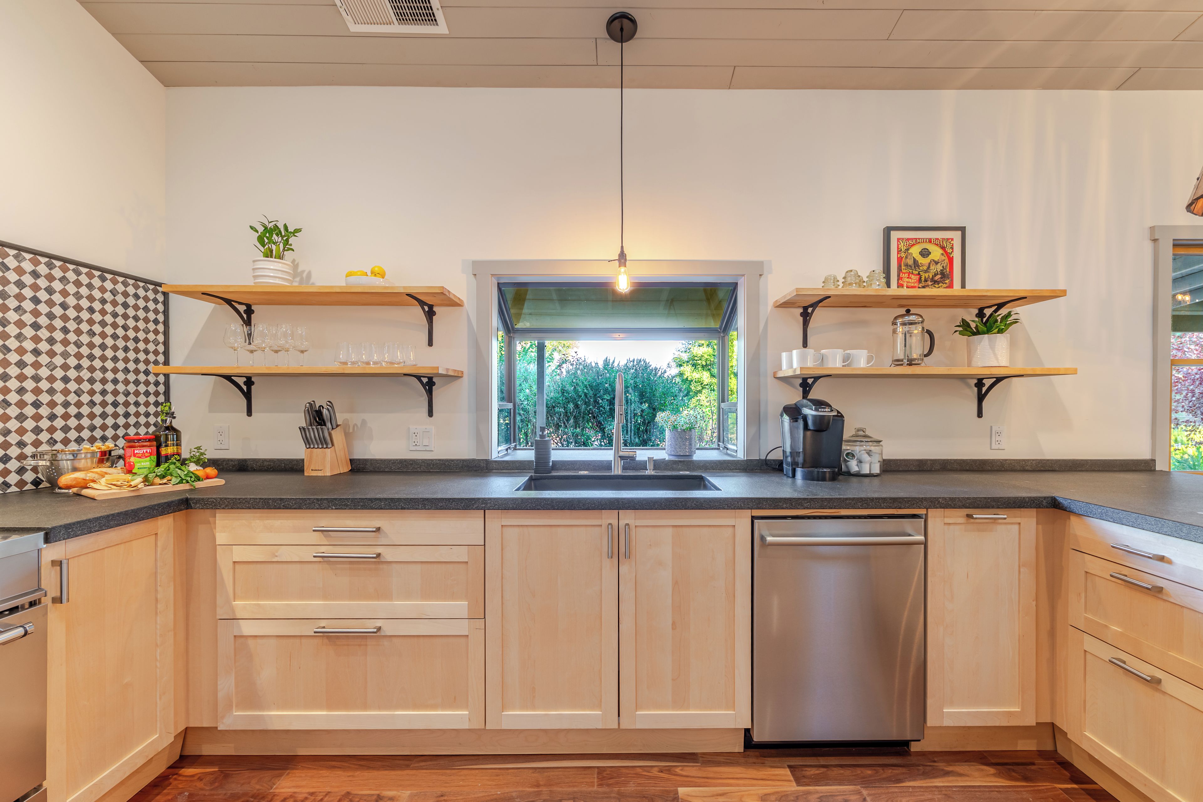 A modern kitchen with a dishwasher, wooden cabinets, a window, and a hanging light fixture.