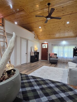 A spacious living room with a wooden ceiling, a ceiling fan, a large stone planter, a checkered rug, and a door leading to another room. - Peaceful mountain cottage near Massanutten, JMU, National Park & Shenandoah Rive (Elkton)