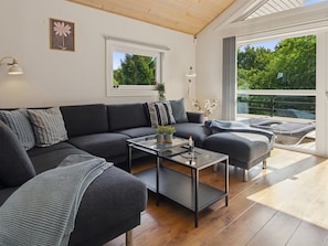 A modern living room with a dark grey sectional sofa, a glass coffee table, and a view of greenery through large windows. - Vacation home "Elle" by Interhome (Toftlund)
