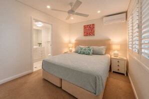 A bedroom with a bed, bedside tables, a ceiling fan, an air conditioning unit, a window with blinds, and a door leading to a bathroom. - Kayannee II - Ocean views, 4 beds, AC (Sunrise Beach)